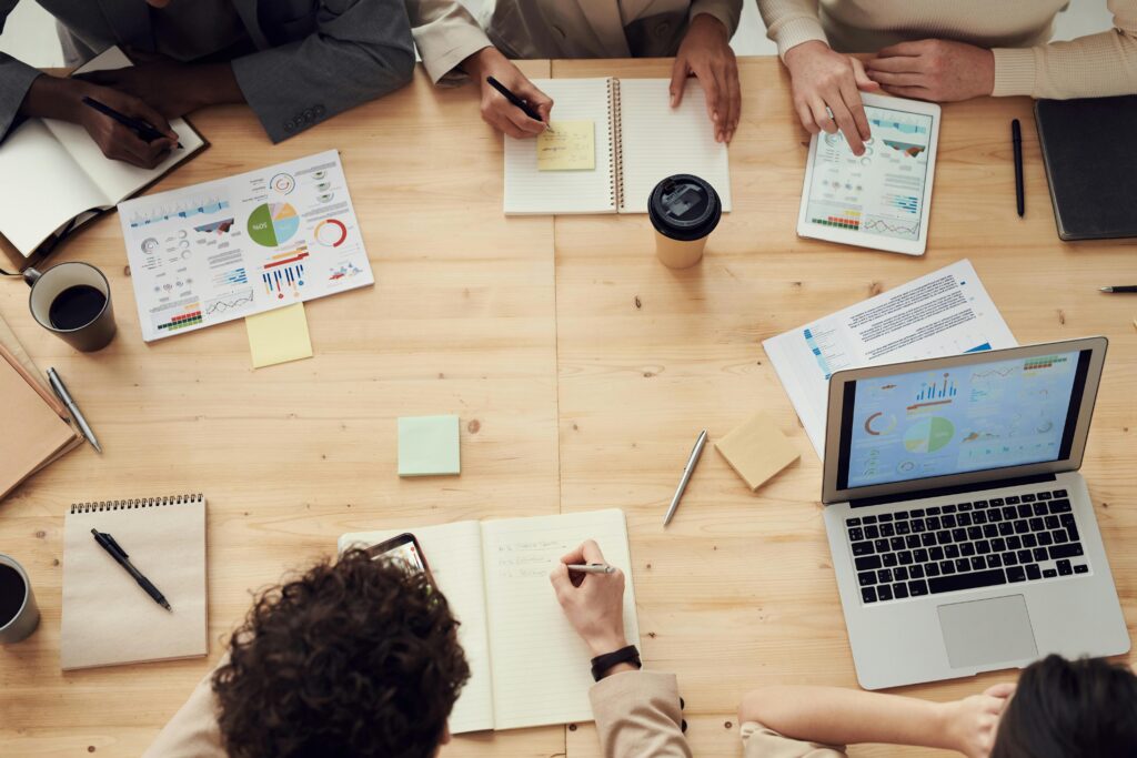 Four colleagues around a table reviewing printed charts, notebooks and a laptop dashboard